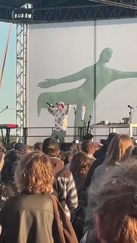 A man wearing a white hoodie with a design on it and a blue baseball cap is performing on a stage in front of a large crowd. He is holding a microphone in his right hand and has his left arm extended to his side. The stage has a large white backdrop with a faint green silhouette of a person on it. There are musical instruments and equipment on the stage. The crowd is diverse in age and gender, with many people looking towards the stage. The lighting suggests it is daytime, possibly late afternoon or early evening. The overall scene is an outdoor concert or festival.