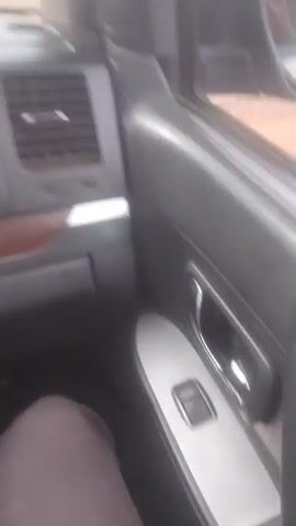 The video begins by showing the interior of a car, focusing on the dashboard, air vent, and door panel. It then transitions to an exterior view, seen from the perspective of the car's passenger side. A side mirror reflects a bustling outdoor scene. A dirt road is visible, populated by numerous people and various vehicles, including motorcycles and what appears to be an auto-rickshaw. Stalls and structures suggest a market or roadside gathering. The scene is active with people walking, standing, and interacting. The sky is overcast. The camera pans slightly, offering glimpses of different individuals and elements within the crowded environment, including the reflection of the car's interior in the side mirror.