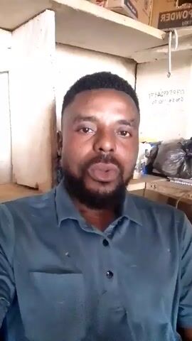 The video shows a man speaking directly to the camera. He is wearing a blue collared shirt. The background appears to be a cluttered room or storage area with shelves, boxes, and bags visible. The man has a beard and short hair. At one point, he covers his eyes with his hand, and at another, he touches his neck and chin. There is text written on the wall behind him.