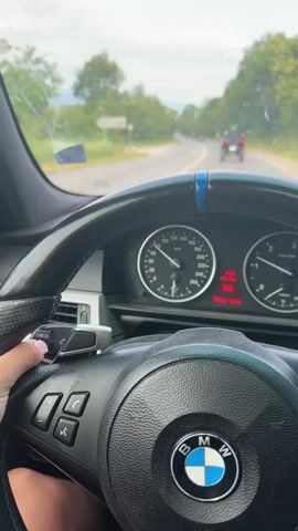 The video shows the interior of a BMW car, with the dashboard and steering wheel clearly visible. The car is moving on a paved road with trees and vegetation on either side. Ahead of the car, a black four-wheeled ATV is traveling in the same direction. The ATV has a rider wearing a helmet. There is another vehicle, a car, visible in the distance ahead of the ATV. The sky is overcast.