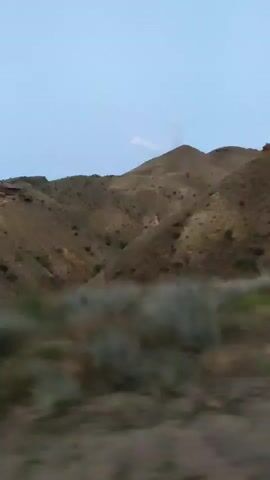 The video shows a view from a moving vehicle looking towards a mountainous landscape. The mountains are covered in sparse, dry vegetation. The sky is a pale blue. The foreground shows a blurred out area that appears to be the side of the road and a guardrail. There are no people or animals visible in the video. The scene appears to be a rural or remote area.