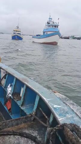 This is a video of boats in the water. In the foreground, the edge of a blue boat is visible, with ropes and buoys attached. In the middle ground, two larger fishing boats are anchored. The boat on the right is white with blue trim and has the name "SANTA ESMERALDA" written on its side. The boat on the left is blue and white with a yellow stripe. The background shows a hazy sky and a distant coastline with more boats and structures, suggesting a port or harbor. The water is choppy, indicating a moderate level of activity. The weather appears overcast and possibly a bit windy. The scene is likely Chimbote, Peru, given the context. No people are clearly visible in the video.