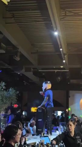 A man with distinctive face paint and brightly colored hair stands on a counter in what appears to be a crowded bar or club. He is wearing a blue shirt and black pants with reflective embellishments. His right arm is raised, and his hand is in a gesture as if addressing the audience. Behind him, a large illuminated sign with the letters "RC" is visible. The ceiling is industrial, with exposed beams and lighting fixtures. Various people are visible in the background, some seated at tables, others standing. In the foreground, several people are visible at a bar, some wearing uniforms that suggest they are staff. Lighting from above and stage lighting cast colorful illumination on the scene.