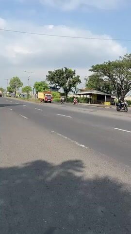 The video shows a bright, sunny day on a paved road in Siring, East Java. The camera is positioned low to the ground, giving a dynamic perspective of the moving vehicles. In the foreground, the asphalt road stretches out, with white lines marking the lanes. 

The main subjects are vehicles. A red and yellow truck with a large black tarp covering its cargo is prominently featured, moving from left to right across the screen. Further down the road, two motorcycles are visible, also heading in the same direction. One motorcycle has a rider wearing a helmet, while the other rider's attire is less clear. In the background, there are trees and some buildings, including a small yellow shelter. The sky is blue with scattered white clouds.

The scene depicts normal daytime traffic. The overall mood is one of everyday activity in a semi-rural or suburban setting. The sunlight suggests it is likely mid-morning or afternoon. There is no discernible text in the video that provides additional context. The camera's movement, tracking the vehicles, creates a sense of motion and immersion in the scene.