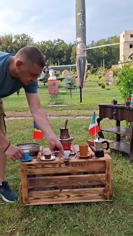 A man is preparing coffee outdoors. He is wearing a blue t-shirt and khaki shorts. He is standing behind a wooden coffee table with a cezve on it. He is holding a small cup in his left hand and a spoon in his right hand. There are two small flags on the table, one Romanian and one Italian. In the background, there is a lawn with trees and a building. There is also a large metallic object that looks like a rocket or missile standing upright. The man pours coffee into two cups.