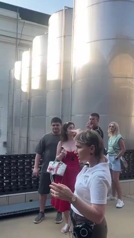 The video shows a group of people standing in front of large, silver cylindrical tanks. The tanks appear to be made of stainless steel and are lined up in a row. A woman in a white shirt is speaking to the group, gesturing with her hands. She is likely a tour guide or an employee of the facility. The group consists of several adults, including men and women. One woman in a red dress is holding a white purse. The environment appears to be outdoors, possibly at a winery or brewery, given the presence of the large tanks, which could be fermentation tanks or storage vessels. The sky is blue and the sun is shining, casting bright reflections on the metal tanks. There is a walkway or platform in the foreground where the group is standing. The overall scene suggests a guided tour or an educational visit to an industrial facility.