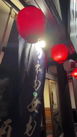 A vertical, low-angle shot captures the exterior of a Japanese establishment at night. Two large red paper lanterns hang prominently, illuminated from within. A dark blue banner with golden Japanese characters is draped to the left, partially obscuring a wooden doorway with glass panels. Further to the right, a string of smaller red and white paper lanterns is visible, also lit. The scene suggests a traditional Japanese restaurant or bar entrance. The lighting creates a warm, inviting glow against the dark night.