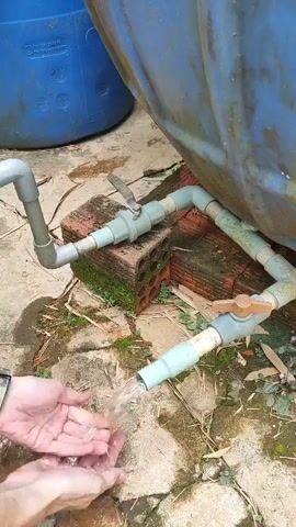 A person is washing their hands under a water pipe that is connected to a large blue plastic tank. The pipe has two valves, both of which are open. The ground is wet and there are some bricks and plant debris visible. The person is wearing a watch on their left wrist and a ring on their left ring finger. The video is filmed from a close-up perspective, focusing on the hands and the water flow. The lighting suggests an outdoor environment.