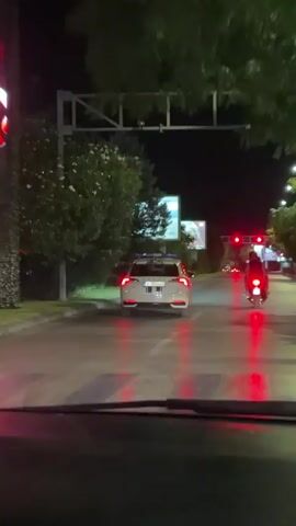 The video, filmed from inside a vehicle at night, shows a white police car with blue and white stripes on its side and rear, and flashing blue lights on its roof, driving down a street. The police car has a license plate that is partially visible and reads "PG MP861". To the right of the police car, a red scooter with a rider wearing a helmet is also visible, with its red taillight illuminated. In front of the police car, traffic lights are red, indicating that vehicles should stop. There are also billboards and trees visible along the side of the road. The street appears to be wet, possibly from rain, as reflections of lights are visible on the asphalt. The perspective suggests the video is taken by a passenger in a vehicle following the police car.