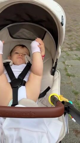 A close-up shot shows a baby in a stroller. The baby is wearing a white onesie and pink socks on its feet. The baby is lying on its back with its legs and arms up in the air. The baby's face is looking towards the camera and it appears to be smiling. The stroller is a light-colored fabric with a brown handle bar. A yellow toy is attached to the handle bar. The background is blurred and appears to be an outdoor setting with a brick walkway. The video is shot from a slightly elevated angle looking down at the baby.