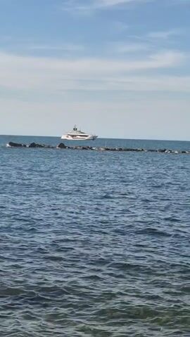 The video shows a wide shot of a body of water, likely the ocean, with a clear sky and scattered clouds. In the background, a large white yacht is visible sailing on the water. In the foreground, there is a line of rocks or breakwater partially submerged in the water. The water itself is blue with gentle ripples. The overall impression is a serene coastal scene.