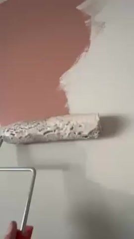 The video shows a person using a paint roller to paint over a wall. The wall is partially covered with a pink paint, and the person is painting over it with a lighter, possibly white, paint. The paint roller has a gray roller cover and a red handle. The person's hand and part of their arm are visible as they move the roller up and down the wall, covering the pink paint with the lighter color. The background is slightly out of focus. The environment appears to be indoors, possibly a room in a house or apartment.