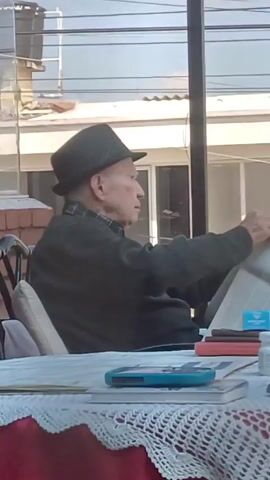 An elderly man is sitting at a table outside, reading a newspaper. He is wearing a dark jacket and a dark hat. The table is covered with a white lace tablecloth. There is a blue cell phone, a red object (possibly a wallet or notepad), a small white bottle, and several books on the table. The man turns the pages of the newspaper as he reads. Behind him is a building with glass windows. There are power lines visible against a light blue sky.