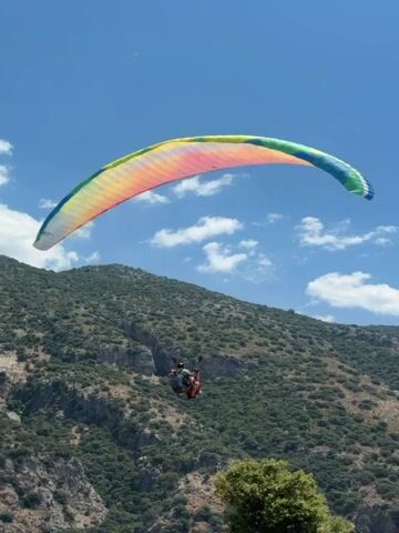 A paraglider is seen in mid-flight against a backdrop of a lush green mountainside and a clear blue sky with scattered clouds. The paraglider, a person suspended in a harness below a colorful, arched parachute, appears to be descending. In the foreground, several people are visible near a grassy area with some trees, and what looks like an outdoor seating area for a restaurant or cafe. Some paragliding equipment is laid out on the grass. The overall scene depicts an outdoor recreational activity in a scenic, mountainous environment.