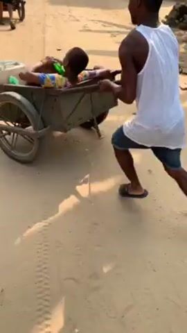 A short video shows three people pushing carts on a dirt road. The person in the front cart wears a white tank top and black shorts and is pushing the cart while the person in the back is riding in it. Another person is pushing a similar cart behind them. Two women run alongside the cart in the foreground. The man in the front cart loses control, and the cart veers off the road into a patch of trees. The man and the person in the cart fall into the foliage.
