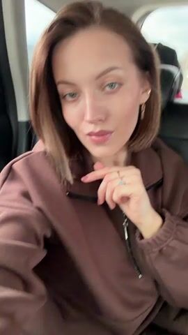 A young woman with short brown hair and light-colored eyes is featured in a selfie video taken from inside a car, likely in the backseat. She is wearing a brown hooded sweatshirt or jacket with a zipper at the neck. On her left hand, she wears a ring with a light-colored stone, and she has chain-link style earrings. Throughout the video, she looks directly at the camera, occasionally adjusting her hair or ear. Her expressions are subtle, ranging from a slight smile to a gentle pout. The car's interior is visible, with dark seats and bags or luggage seen in the upper right background through the window. The exterior views through the car windows show an overcast sky and glimpses of trees and a building with a red roof, suggesting a daytime setting in a rural or suburban area. The scene is set in Velmogovo, Russia.