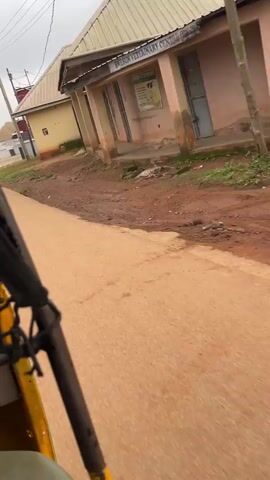 The video shows a point-of-view shot from a moving vehicle, likely a rickshaw, with the right side of the vehicle visible in the foreground. The vehicle is traveling on a dirt and asphalt road. On the right side of the road, there are several buildings. The first building visible is a light-colored structure with a blue sign that reads "BWESUN VETERINARY". Further down the road, a red and white building with the sign "REDTECH MEDICAL SERVICES" is visible. There are also signs for a church. The ground is a mix of dirt and some puddles, indicating it may have rained recently. The sky is overcast.