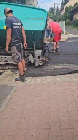 A construction worker in a dark t-shirt and grey shorts stands next to a large, green asphalt paving machine. He appears to be holding a cigarette in his left hand. Two other construction workers are visible behind the machine. One, wearing a red t-shirt and red shorts, is using a tool to work on the asphalt. The other, wearing a blue shirt and jeans, is also behind the machine. The asphalt paving machine is actively laying down a fresh layer of asphalt on a road. The surrounding environment appears to be a residential street with houses visible in the background. The ground consists of a paved sidewalk and the newly laid asphalt road.