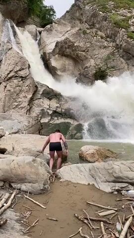 A man stands on the rocky shore of a river, facing a large, powerful waterfall. He is wearing black swim trunks and has tattoos on his back. The waterfall cascades down a steep, rocky cliff face, creating a large spray of mist. The river is murky brown and littered with debris, including branches and plastic bottles. The surrounding environment is rugged and natural, with lush green vegetation growing on the cliffs. The sky is overcast.