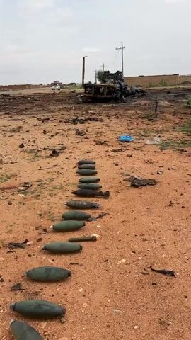 The video shows a desolate, dusty, and barren landscape. In the foreground, a line of mortar rounds, mostly green with some dark colored ones mixed in, are laid out on the dirt. In the background, a destroyed and burnt-out vehicle is visible, with debris scattered around it. A utility pole stands behind the vehicle. The sky is overcast.