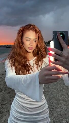 A person with shoulder-length wavy red hair, wearing a white sheer dress, poses on a sandy beach. The person looks off to the right and seems to be posing for a photo or video, as a second person holds up a rectangular light source directly in front of them. The sky behind them shows a dramatic sunset with dark, cloudy formations and a pink and orange glow on the horizon. The beach is lined by the ocean, with small waves visible in the background.