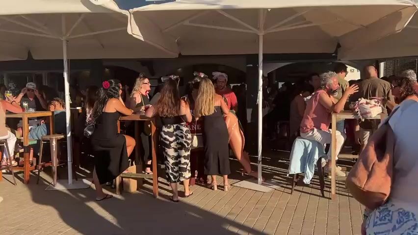 The video shows a crowded outdoor patio area of a bar or restaurant, with tables and umbrellas providing shade. Numerous people are seated and standing, engaged in conversation and drinking. Several women are prominently featured, dressed in what appear to be party or evening attire. One woman in a black dress is leaning on a table. Another woman in a black dress and a woman in a cow-print skirt are interacting near the center of the frame. A person in a pink shirt is seated on the right side, and a man in a white shirt and shorts is visible on the left. The ground is paved with interlocking bricks. The overall atmosphere suggests a social gathering during daylight hours, possibly late afternoon or early evening given the lighting and shadows.