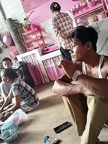 The video shows an indoor scene with several people. In the foreground on the right, a man in a white tank top and tan pants is sitting and looking to the left. He is wearing a watch on his left wrist. In front of him on the floor are a cell phone and a pack of cigarettes. To the left of the man, an older woman in a checkered shirt and patterned skirt is sitting on the floor. A plastic bag and a woven basket are near her. Behind them, another person is sitting down. In the background, a person in a plaid shirt is standing and facing away from the camera. There is pink shelving unit with items on it in the background, and pink curtains are visible. The room appears to be sparsely furnished with a concrete floor.