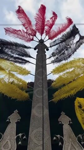 This is a medium shot, captured from a low angle, of an ornate, metallic structure adorned with colorful feathers. The structure is tall and slender, with intricate carvings and Arabic script covering its surface. Surrounding the central structure, several ornate metallic objects with hanging bells are visible, two prominent ones flanking the base of the central piece.

The structure is decorated with clusters of large, fluffy feathers in shades of red, black, and yellow. The red and black feathers are positioned higher up, fanning out from the top of the structure, while the yellow feathers form a vibrant halo around the mid-section. The metallic elements appear to be silver or polished steel, and the craftsmanship suggests a ceremonial or religious context.

The scene is lit by natural light, suggesting it is daytime. The sky is visible in the background, appearing partly cloudy. The overall impression is one of solemnity and grandeur, possibly indicating a religious procession or commemoration, given the elaborate decorations and script. The presence of bells also suggests a ceremonial event. The location is identified as Babol, Iran, which aligns with the style of the artwork and script. The visual elements do not explicitly depict people, but the scale and decoration imply human involvement in its creation and use.