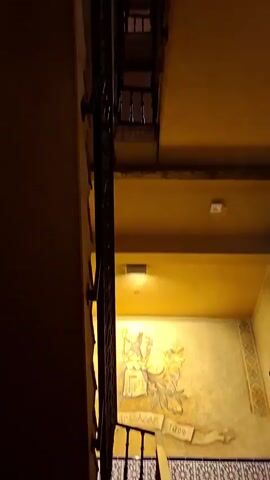 The video shows a view looking down a staircase. The staircase has dark metal railings and appears to be made of wood or stone steps. The walls are painted a warm yellow or ochre color, and there is a mural visible on one of the walls. The lighting is dim, with some light fixtures visible on the ceiling. The camera angle is from above, looking downwards, creating a sense of depth and perspective.