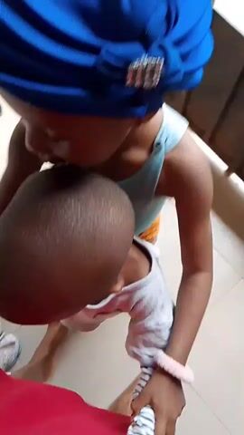 This video shows a young child wearing a blue headwrap with a jeweled accent, and a light blue top. The child is holding and interacting with a baby. The background appears to be indoors with tiled flooring and wooden structures visible. The interaction is brief and primarily focused on the two children.