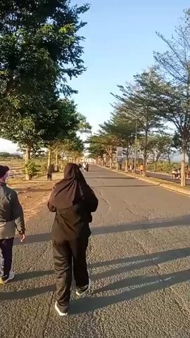 A person walks down a paved road lined with trees on a sunny day. The person is wearing a black hijab and black pants. Another person is walking in front of them to the left, wearing a gray jacket and dark pants. There are other people and vehicles in the distance on the road, and signs are posted along the side. The sky is clear and blue.