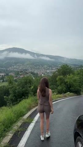 A person with long, dark hair, wearing a brown sleeveless top, brown shorts, and white sneakers, walks away from the camera on a wet, paved road. A white solid line marks the edge of the road on the left, next to green grass and trees. In the background, a town with numerous buildings is visible in a valley, surrounded by green trees. Beyond the town, hills and mountains are covered in mist or low clouds under an overcast sky. A dark vehicle is partially visible on the right side of the road.