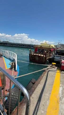 A video shows a view from the deck of a ferry boat. The boat is docked next to a concrete pier. In the background, another boat with a decorative roof is docked. The water is blue, and the sky is clear with a few clouds. A man stands on the deck of the ferry, looking out at the water. The ferry has a white cabin and a blue deck. The pier has a yellow line painted along its edge.