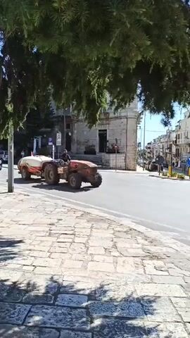 A person is operating a red tractor with a white tank trailer attached, driving down a street. The street has a cobblestone sidewalk on the left and paved road with cars on the right. Trees with green leaves hang over the scene from the top left. Buildings line the street in the background.
