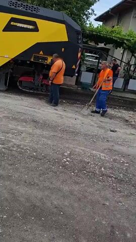 The video features a large yellow road milling machine, bearing the "BOMAG" logo, actively engaged in road construction work. A man in an orange shirt is standing near the machine, while another man on the machine operates it. The milling machine is connected to a white pipe or conveyor belt, through which it appears to be transferring excavated material into the back of an orange dump truck. The dump truck is slowly backing up, aligning itself to receive the material from the milling machine. The ground in the foreground is unpaved and looks like a construction site. Green trees and a building are visible in the background, suggesting an outdoor urban or suburban setting.