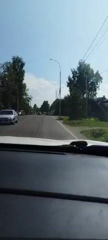 The video shows a view from inside a vehicle driving on a paved road on a sunny day. The road is lined with trees and green vegetation. There are other vehicles on the road, including cars and SUVs. There are also utility poles and wires visible along the side of the road. The vehicle appears to be moving at a moderate speed, and the road ahead curves to the right. There are no pedestrians clearly visible in the frame. The dashboard of the vehicle is visible in the foreground.