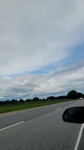 The video shows a view from inside a vehicle traveling on a paved road. On the left is the side of a black minivan. On the right, the side mirror of the camera vehicle is visible. Several other vehicles are on the road, including a white van with writing on its back, a silver car, and another white van. The road is bordered by a grassy field and trees in the distance. The sky is cloudy.