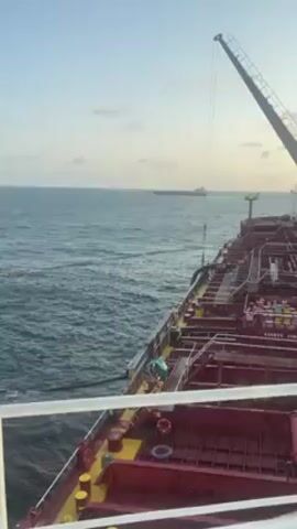 The video presents a view from the deck of a large red tanker ship. The camera is stationary and looks out over the side of the ship. The sea is visible in the background, with another large ship on the horizon. A crane is visible on the deck of the tanker, with a cable extending downwards. The sky is a light blue color. No people are clearly visible. The tanker appears to be on the open ocean.