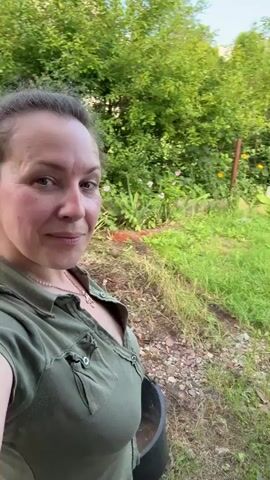 A woman in a green shirt is filming herself outdoors with a dog in the background. The dog is tan and appears to be sniffing the ground. The environment is a wooded area with trees and bushes. There is also a large log in the background. The woman smiles at the camera and the dog moves around.