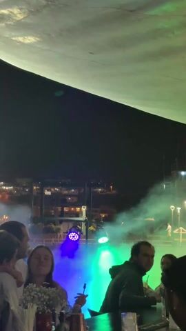 The video captures a group of people at an outdoor gathering at night, possibly a bar or restaurant. They are standing or sitting near a table, against the backdrop of a cityscape illuminated with streetlights and buildings. Stage lights emanate colorful beams and smoke into the air. Some of the individuals are interacting, including a couple who are kissing, while others seem to be enjoying the music.