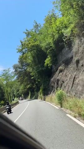 A car drives along a winding road flanked by lush green trees and a rocky incline. The sun shines brightly in a clear blue sky. The road has a white lane marking and a barrier on the left. Another vehicle is visible in the distance ahead.
