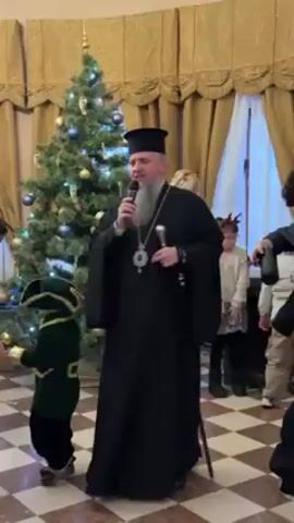 A man in a black cassock, with a white beard and a black hat, speaks into a microphone while standing in front of a Christmas tree. He holds a silver cane in his left hand. Children dressed in costumes are milling around him. A person with blonde hair is visible behind him. The room has beige walls and a patterned floor. Curtains hang in the background.