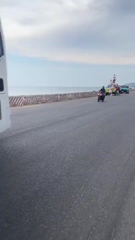 The video shows a scene next to a beach road with a motorcyclist passing by. A yellow off-road vehicle has several people standing around it and on its roof. They are posing and taking photos. A green off-road vehicle is parked further down the road near the beach. The sky is cloudy, and the sea is visible in the background.
