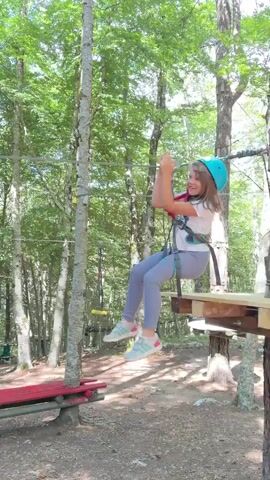 A young girl, wearing a white shirt, gray leggings, and a blue helmet, glides on a zip line through a forest adventure course. She smiles at the camera as she starts, then pushes off a wooden platform, propelling herself along the wire between trees. She lands on another wooden platform, where a person in a red shirt and green shorts is waiting. The trees in the background are green and lush, indicating a summer or spring setting.
