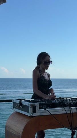 A woman wearing headphones and sunglasses stands in front of DJ equipment on a wooden counter against a backdrop of ocean and blue sky. She adjusts her sunglasses.