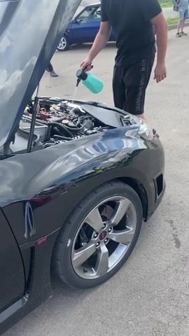 The video depicts a sunny day at an outdoor event, likely a car gathering or race track, as suggested by the presence of various vehicles and a race track in the background. The foreground features a black Subaru, possibly a performance model given the "STI" badge visible on the fender. The hood of the Subaru is open, revealing its engine bay.

A man, dressed in a black t-shirt and shorts, is actively engaged with the car's engine. He holds a light blue spray bottle and appears to be spraying a liquid into the engine compartment. His posture and focused expression suggest he is performing some sort of maintenance or preparation on the vehicle.

In the middle ground, other individuals are visible, some observing the scene. A man holding a black umbrella stands to the right of the Subaru. A dark-colored sedan with the license plate "O427PP 750" is parked behind the Subaru. In the distance, more cars and people are present, with a clear view of a race track and distant hills under a cloudy sky. The overall atmosphere seems casual and focused on automotive activities.

The visible text includes "STI" on the car's fender and "NIKE STRENGTHS" on the man's shorts. The license plate of the sedan is "O427PP 750". The location context is Svetloe Pole, Russia.