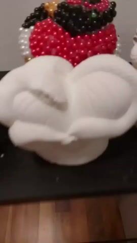 A white sculpture of a snake coiled around a rounded object is on a dark surface. Behind it, out of focus, is a decorative item made of red, black, and white beads in the shape of a bird. The camera pans from left to right, showing a portion of another white object to the left of the snake sculpture.