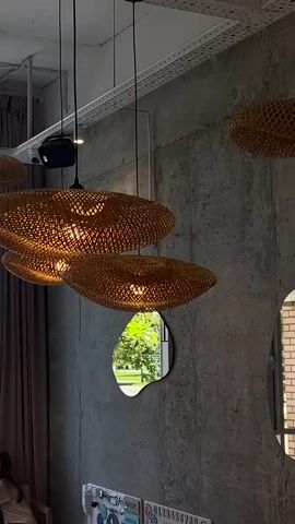 The video shows three woven basket-like pendant lights hanging from the ceiling. The lights are illuminated, casting a warm glow. The walls appear to be concrete, with some texture. Two irregularly shaped mirrors are mounted on the wall. Through one mirror, a glimpse of greenery and trees is visible, suggesting an outdoor scene. Through the other mirror, a brick wall and a window are visible. A brown curtain is partially visible on the left side of the frame. A metal ceiling fixture with a light is also visible at the top of the frame.