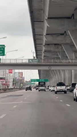 A vertical, low-angle, outdoor video shows a view from a vehicle driving on a multi-lane highway under an elevated highway. The sky is overcast and grey. On the left side of the lower highway, there are shops and billboards. On the right, there are many concrete pillars supporting the elevated highway. Several cars are driving on the highway, including white SUVs, a white truck, and a tuk-tuk. Green signs on the elevated highway indicate directions. The video shows the same scene from beginning to end.