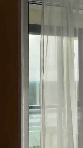 The video shows a view from indoors looking out a window with white translucent curtains. A portion of a beige wall is visible on the left. Outside the window, one can see a distant cityscape with green trees. The bright sunlight causes a lens flare at the top of the frame.